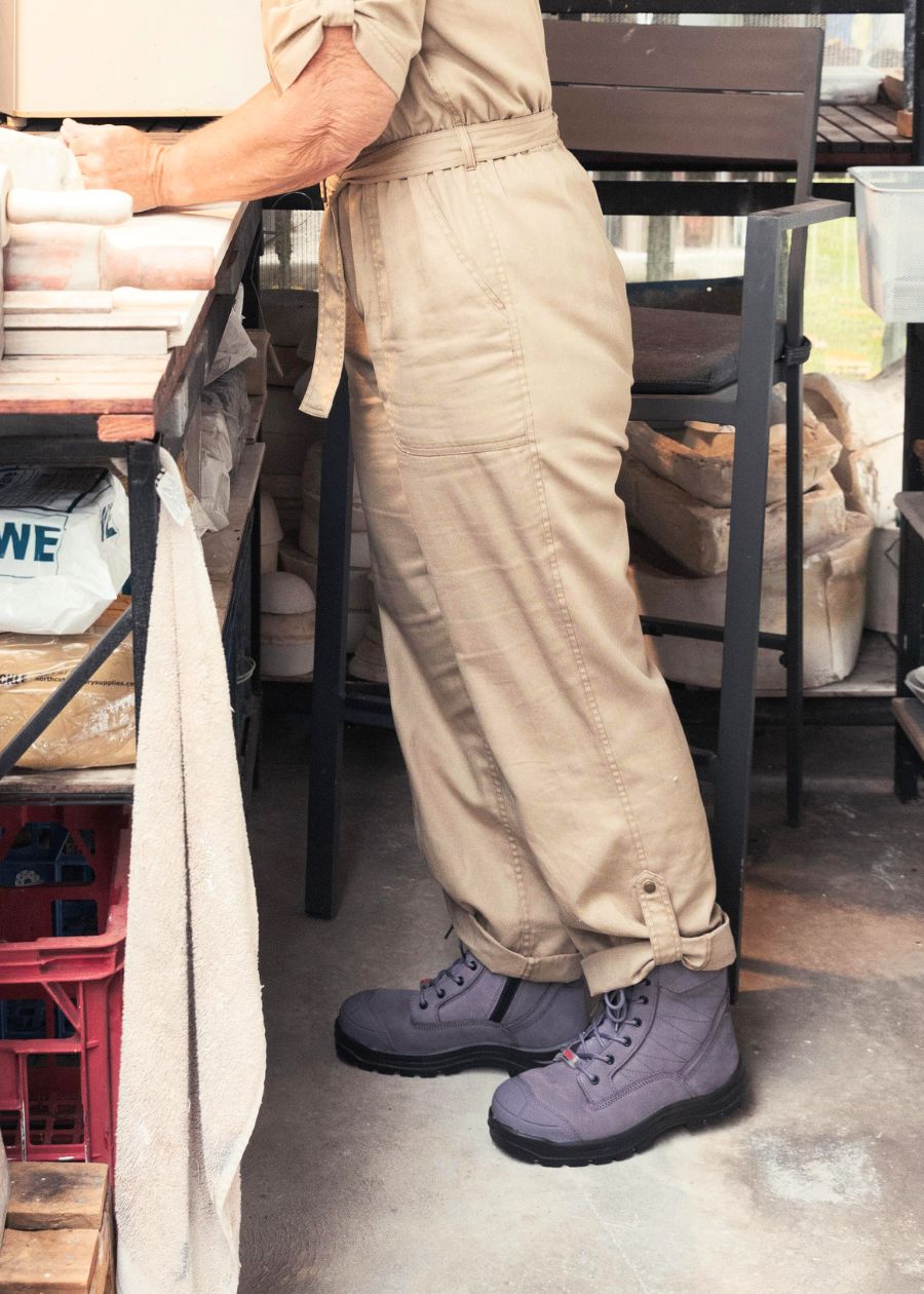 Woman standing in pottery studio in tan jumpsuit and purple lace-up safety boots.