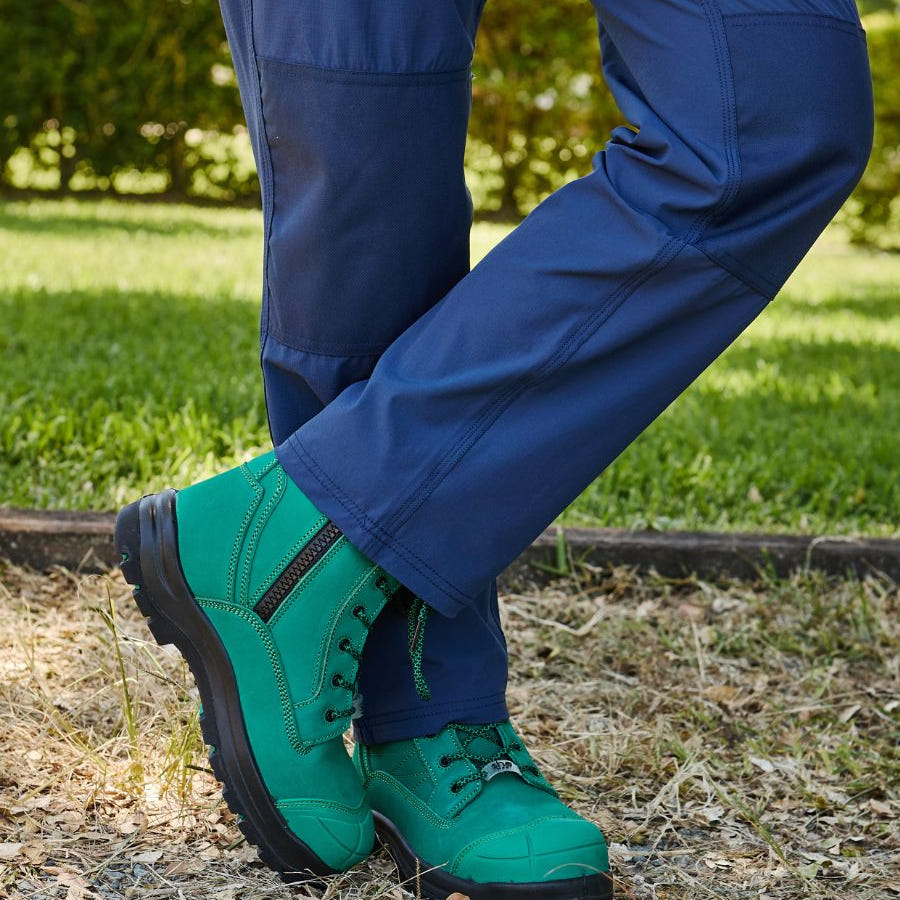 Standing in garden legs crossed wearing long blue workpants, and green zip-up work boots.