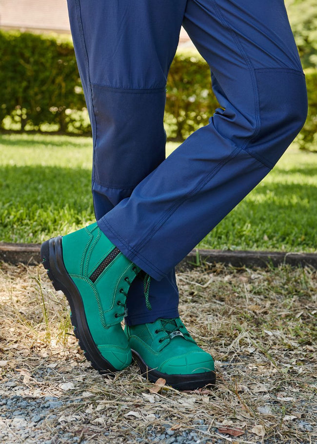 Standing in garden legs crossed wearing long blue workpants, and green zip-up work boots.