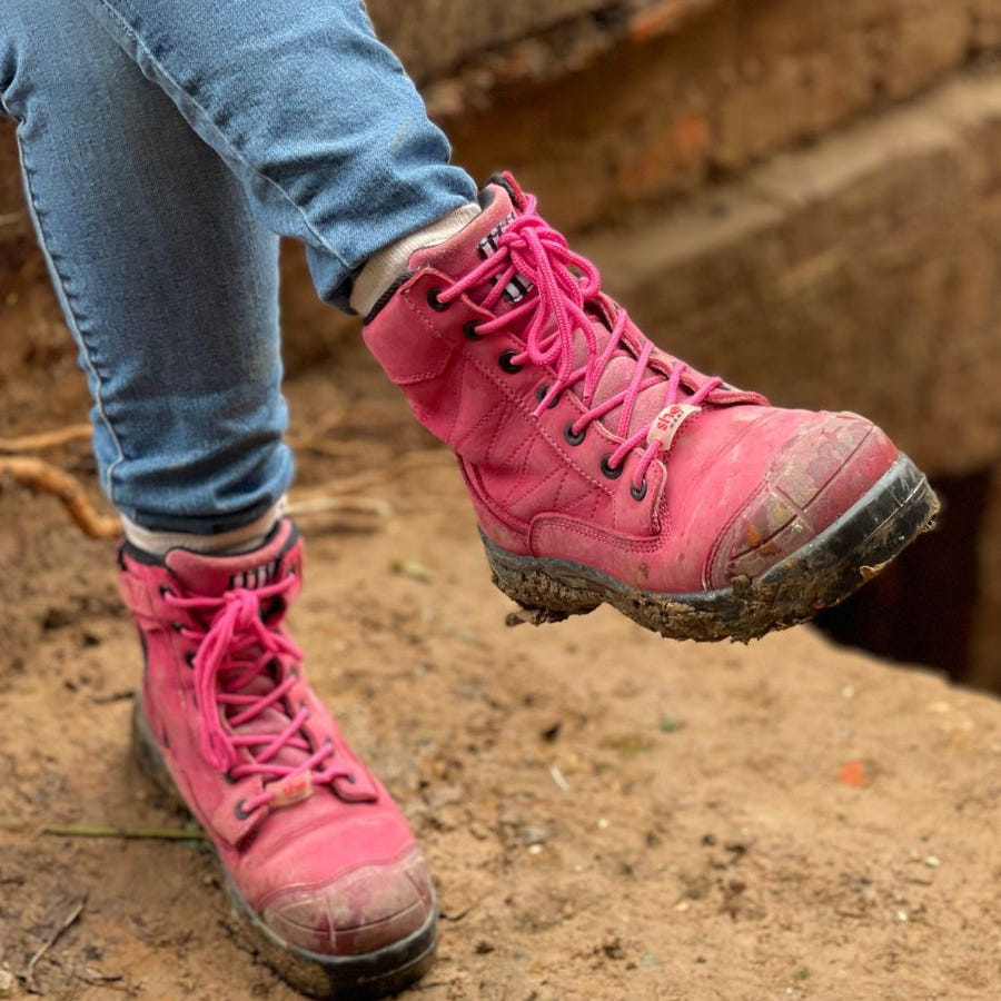 Person sitting on worksite in pink lace-up composite work boots, covered in mud.