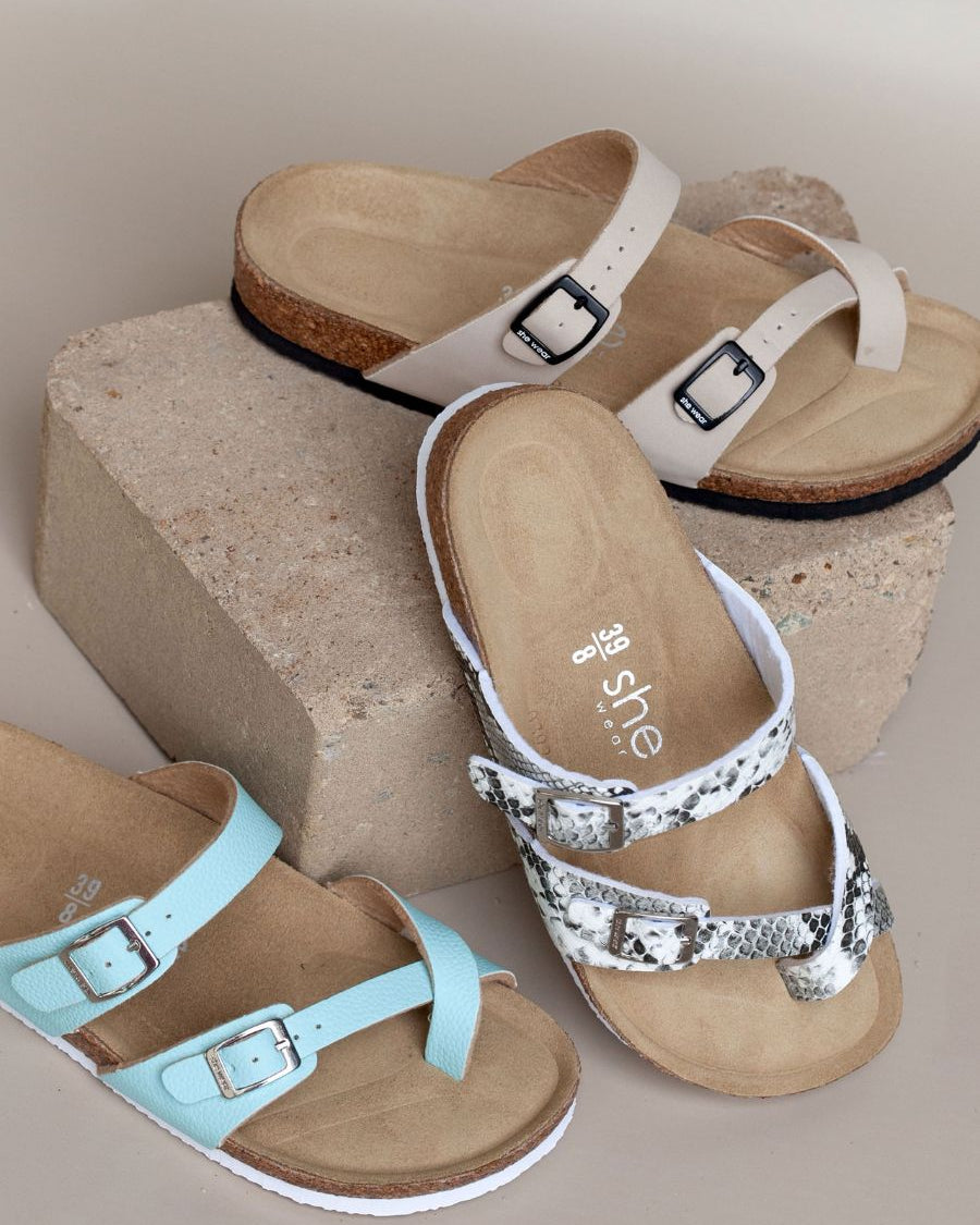 three cork leather slides rest on a brick, showing contoured footbeds and adjustable straps in blue, tan, and a black and white snakeskin print