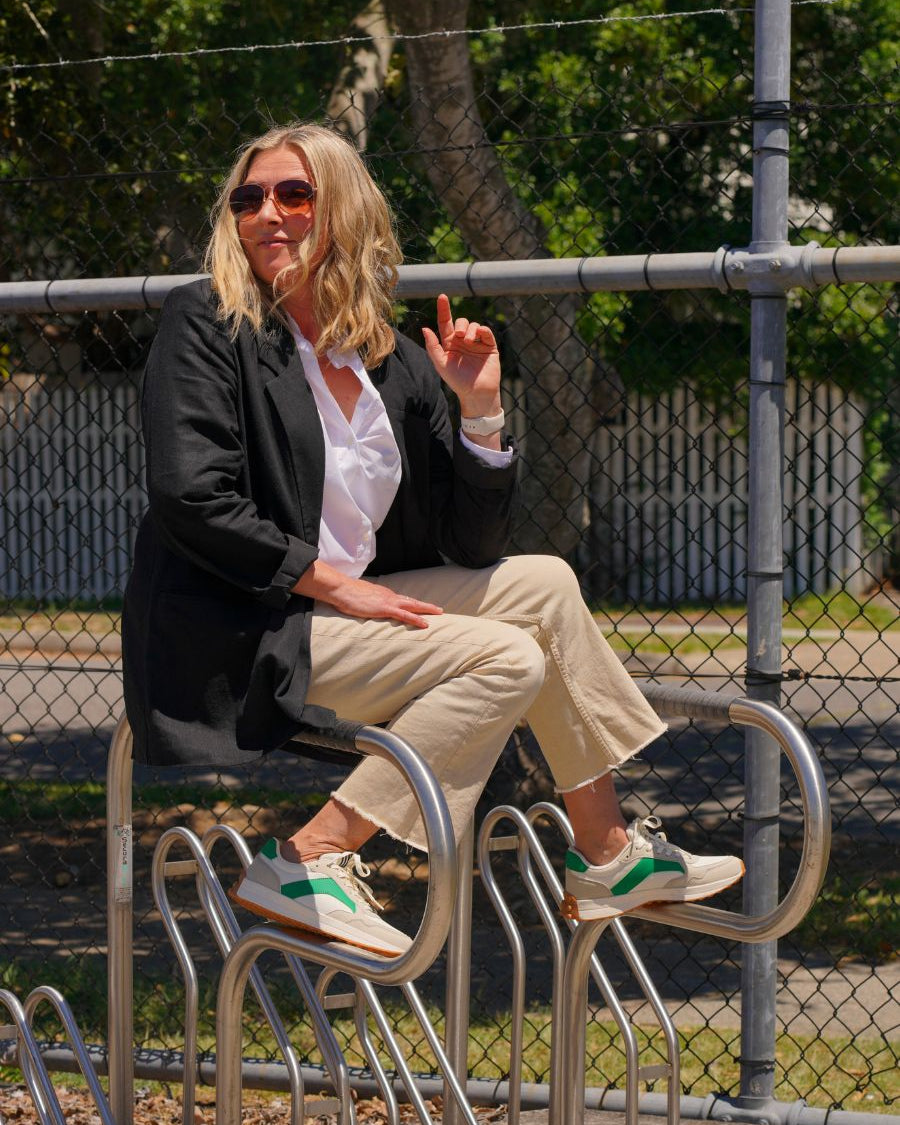 A woman sits on a bike rack wearing a black blazer, tan jeans, and tan and green sneakers
