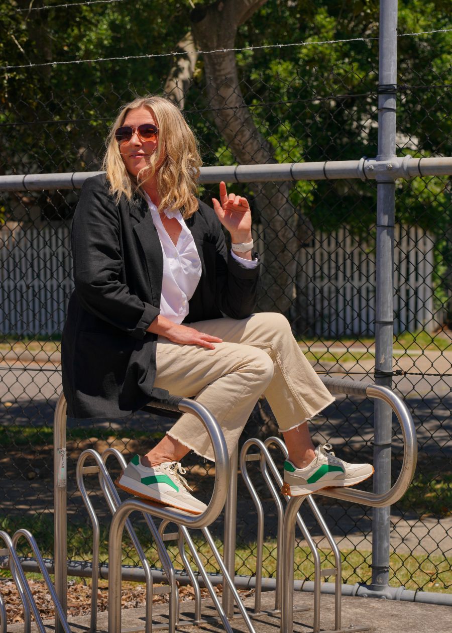 A woman sits on a bike rack wearing a black blazer, tan jeans, and tan and green sneakers