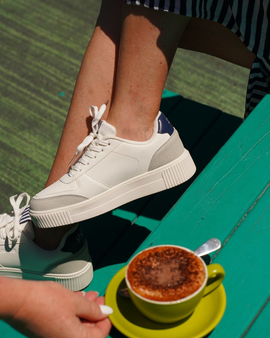 White, navy and grey platform sneakers with a padded heel collar for comfort shown behind a cup of coffee