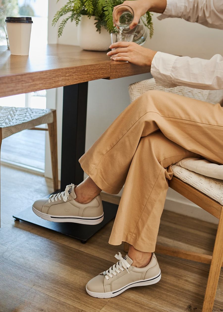 Women sitting at wooden table, with neutral colour tones, wearing stone coloured support sneakers.