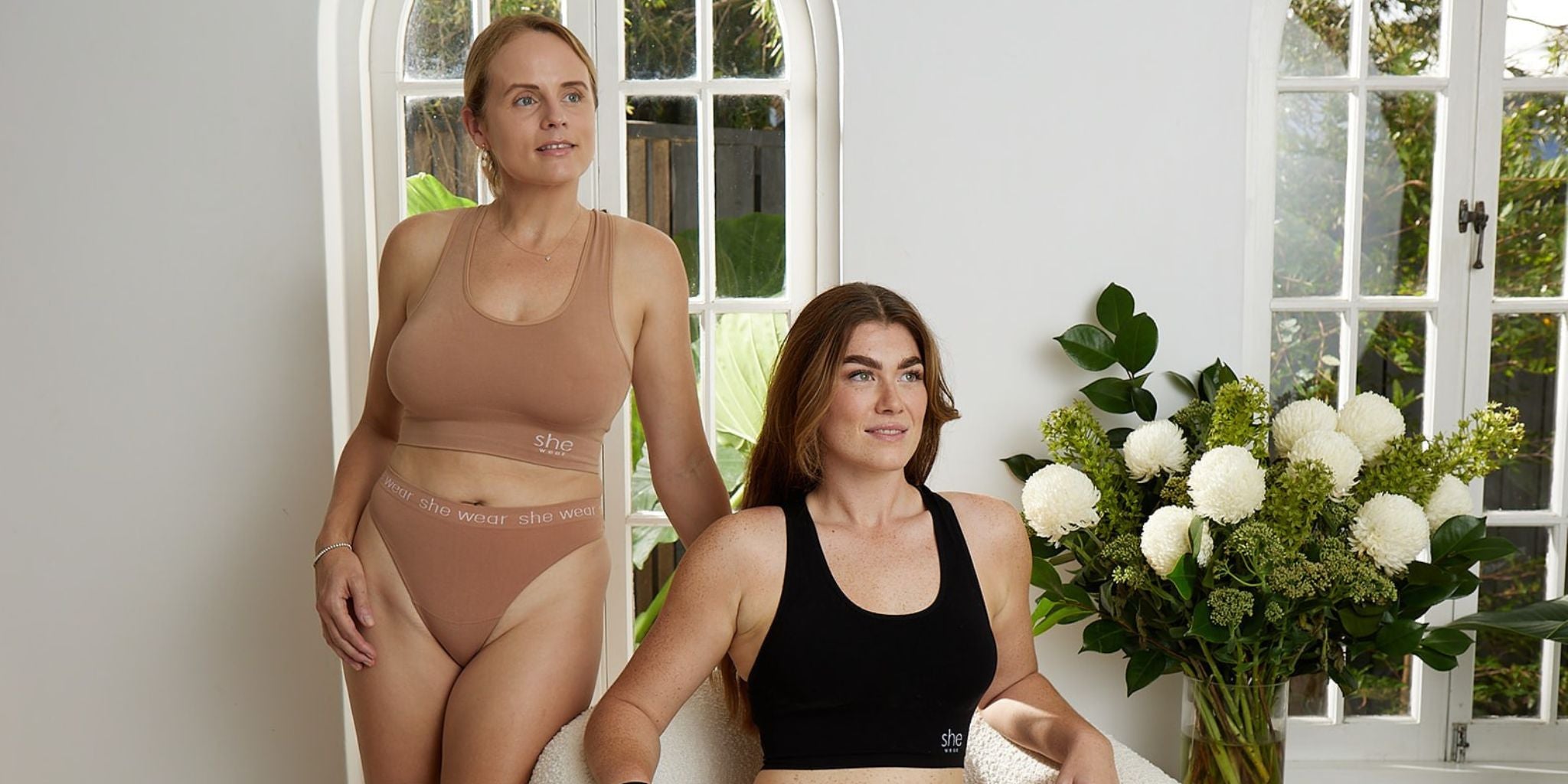 Woman sitting in a black bamboo crop bra with woman on left standing in nude colour bamboo bra and g-string. 