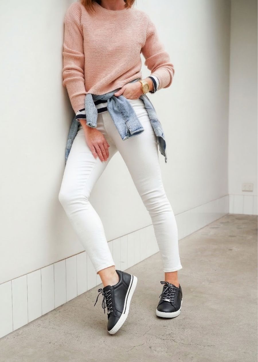 Woman leaning against a white wall wearing black and white supportive sneakers. 